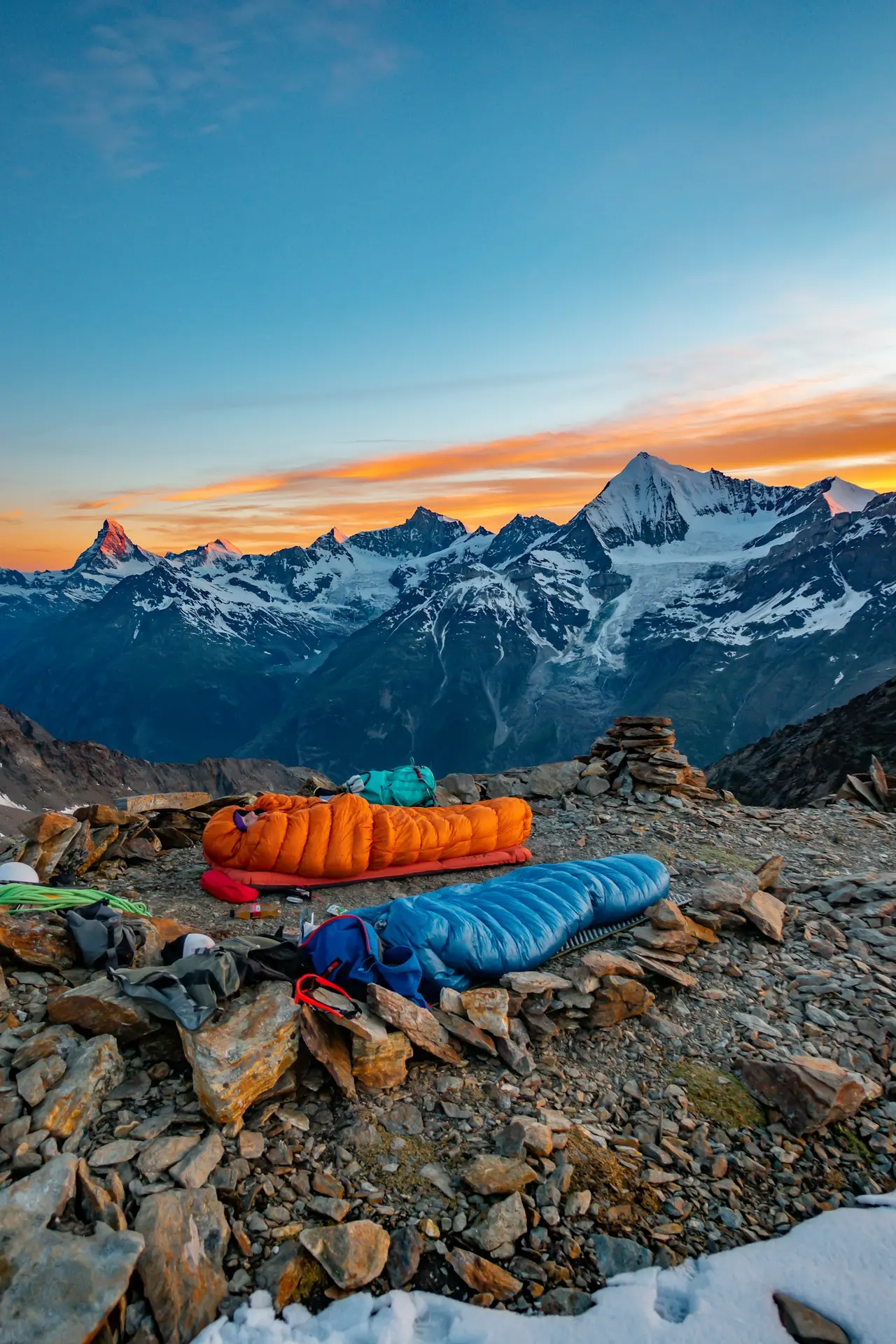 Two down mummy bags laid out on a rocky alpine ridge at dawn: an orange one with its hood drawn up around a sleeper's face, on a thick red inflatable pad, and a blue one beside it on a thin ridged foam mat, snow-covered peaks behind.