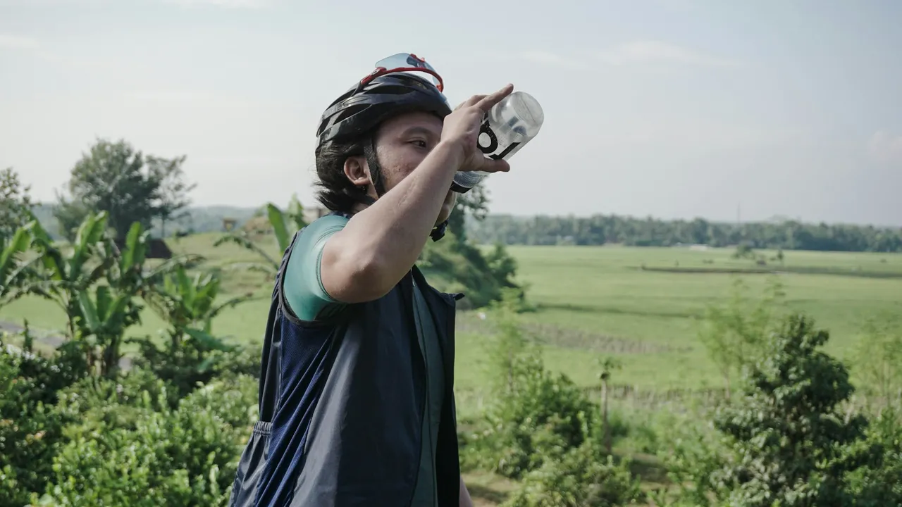 A rider tipping a bottle back to drink, open green fields behind.