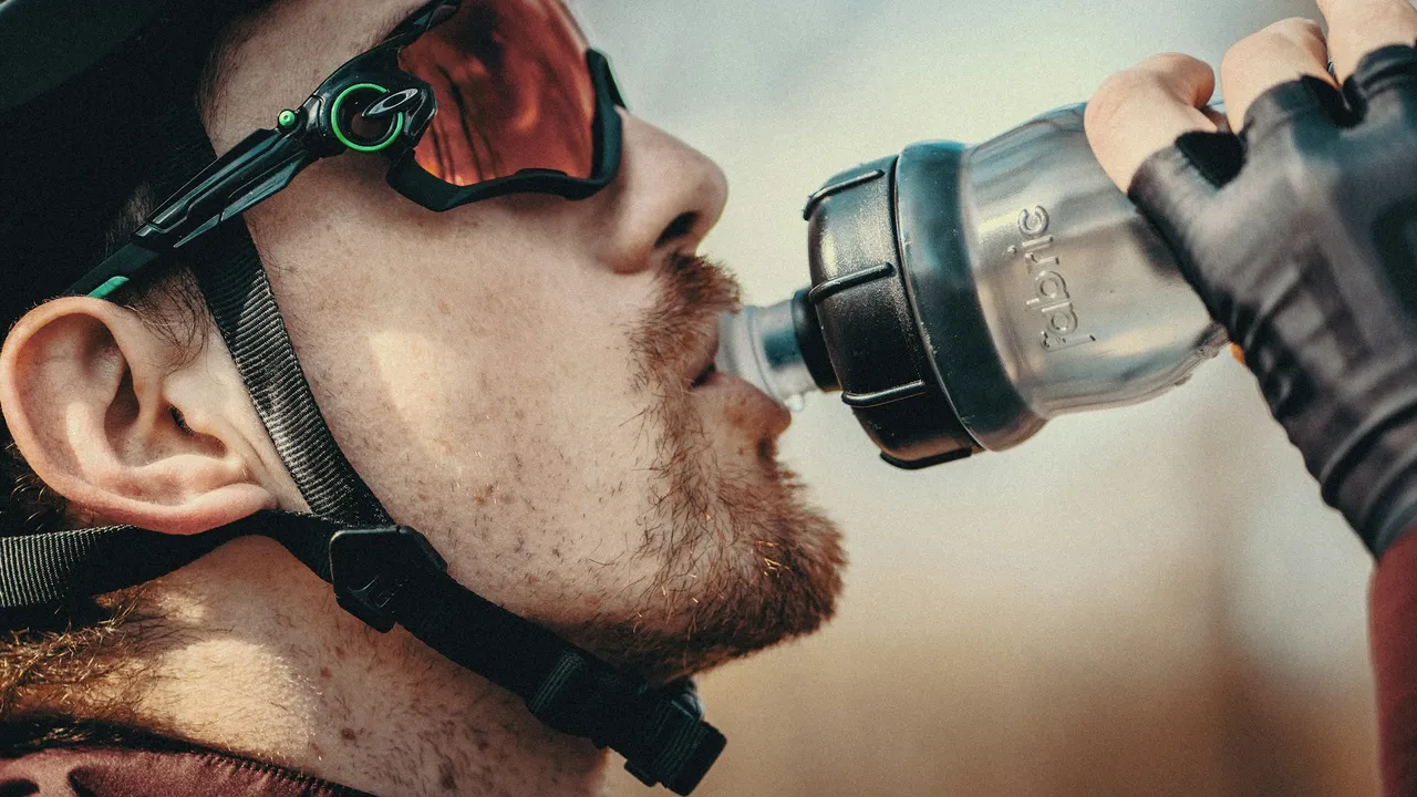 A helmeted rider in sunglasses drinking from a bidon, seen in close profile.
