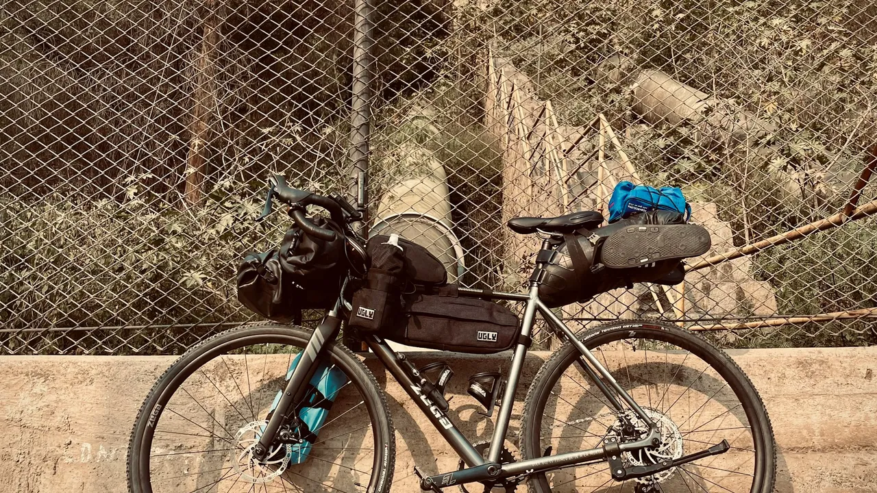 A gravel bike loaded with a bar roll, frame bag and seat pack, leaning against a wire fence.