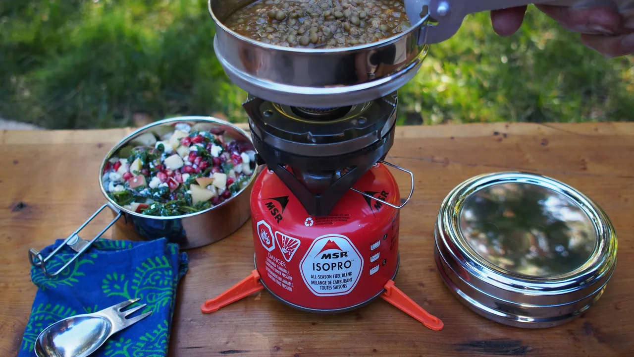 A canister stove on a wooden table with a pot being lifted off it, a bowl of grain salad and a spork alongside.