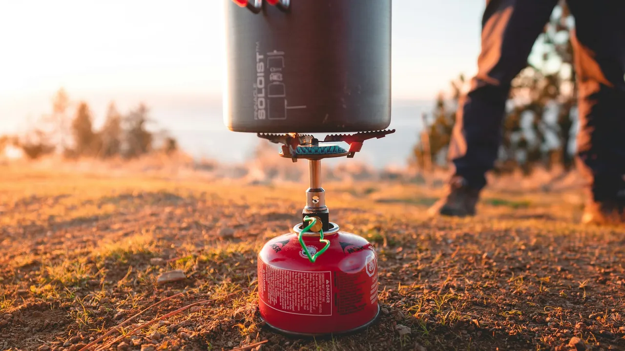 A pot balanced on a canister stove on rocky ground at sunset, a person standing behind.