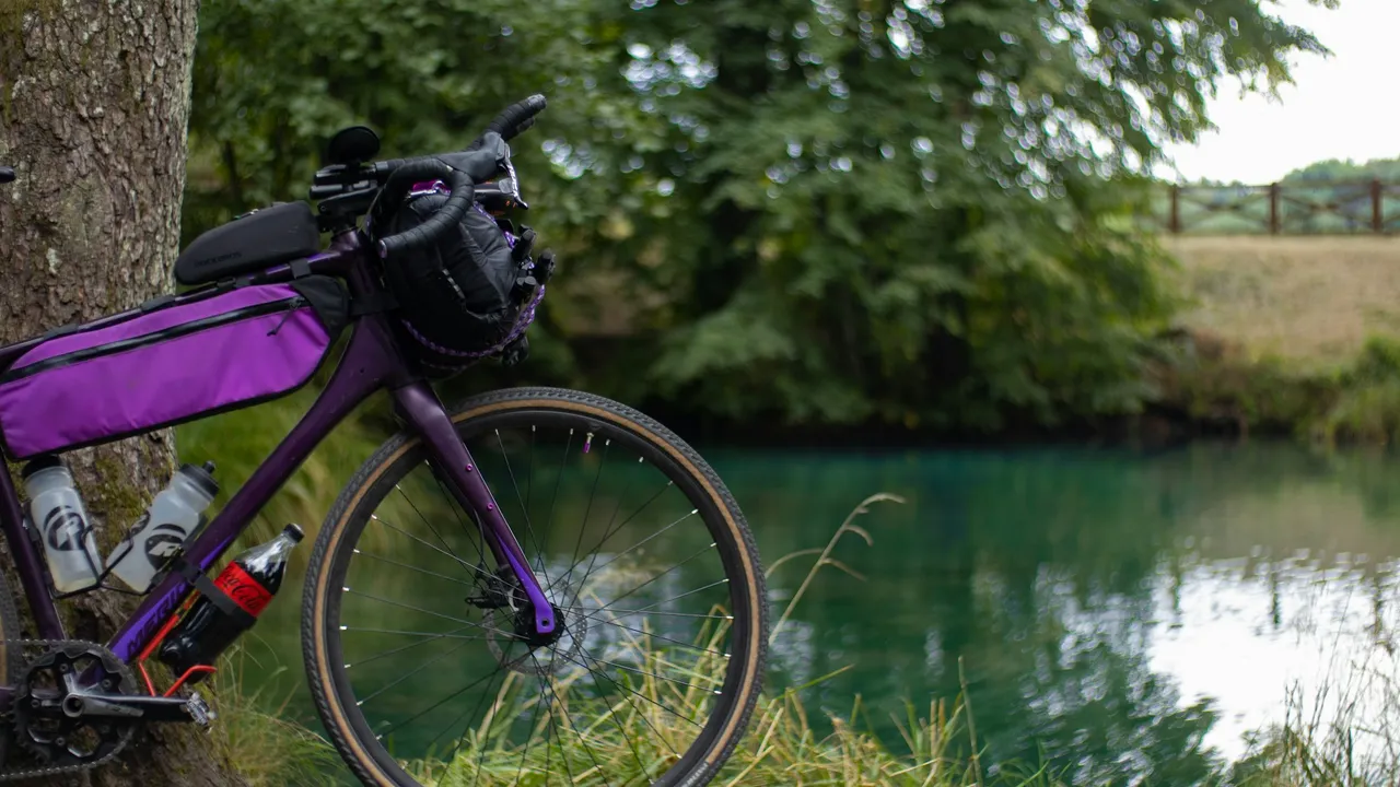 A purple gravel bike with a bar bag and bottles in the down tube cages, parked beside a river.