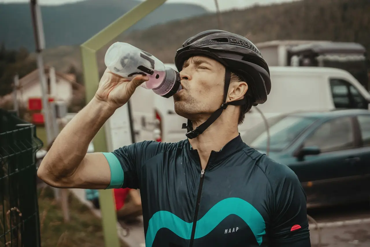 A rider in a helmet drinking from a bidon at the roadside.