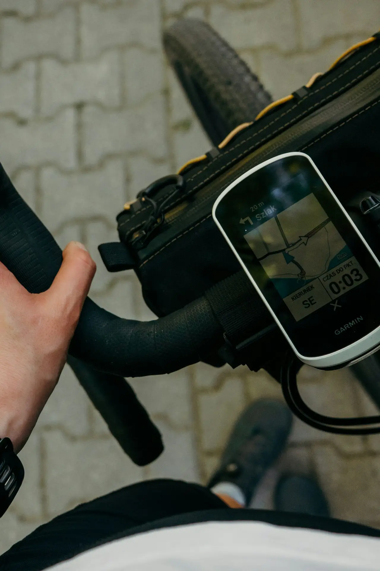 Looking down at a Garmin head unit mounted on a drop-bar gravel bike with a bar bag, its screen showing a map with the route line and the next turn called out at the top, the rider's hand on the bar.