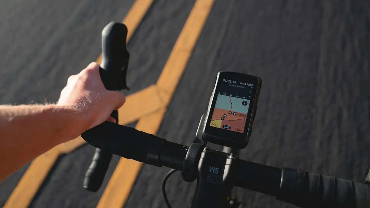 A GPS head unit mounted ahead of the stem showing a route map, the rider's hand on the drop-bar hood and road markings passing below.