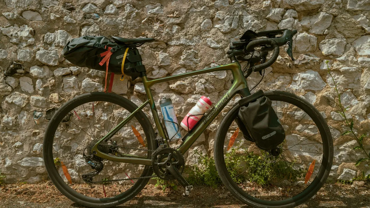An olive gravel bike side-on against a stone wall, a bar bag at the front and a bottle in the down tube cage, the main triangle open.