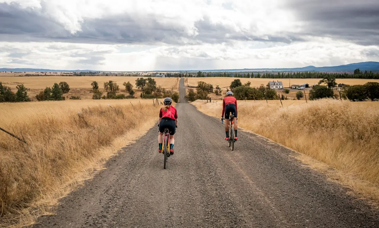 Two riders on loaded bikes on a gravel road running between open fields.