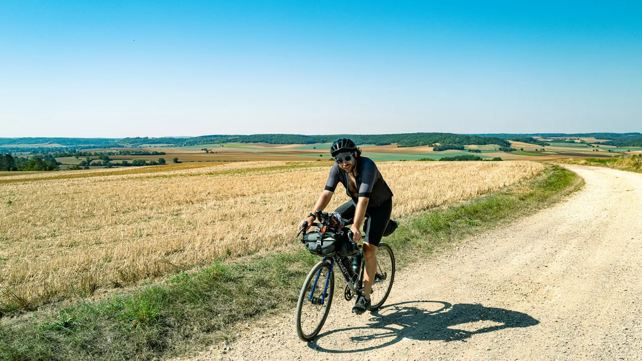 A rider on a loaded gravel bike climbing a chalk farm track between harvested fields under an empty blue sky.