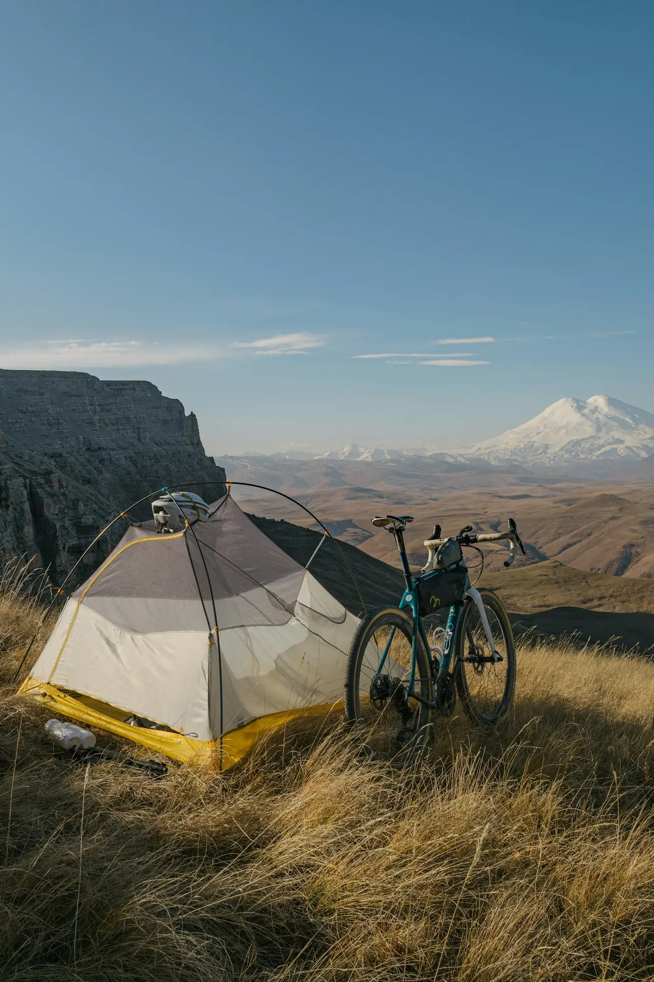 A gravel bike leaning beside a tent on a grassy plateau, carrying a part-length frame bag slung under the top tube and a small bag at the handlebars.