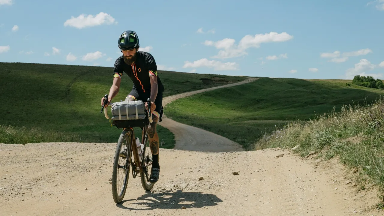 A rider on a loaded gravel bike with a bar roll, climbing a dirt road through open hills.
