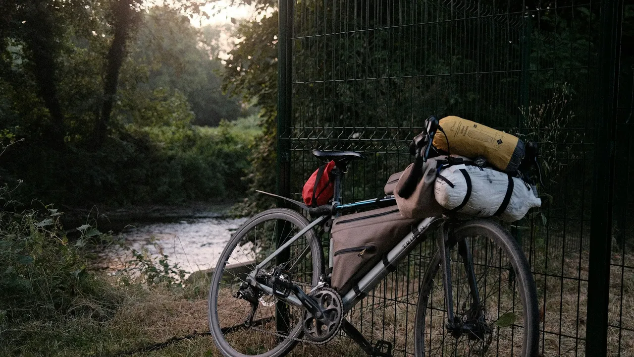 A silver Decathlon Triban drop-bar bike loaded with a frame bag, a bar roll with a white bundle and a yellow stuff sack strapped on, and a small red bag under the saddle, leaning on a mesh fence beside a river at dusk.