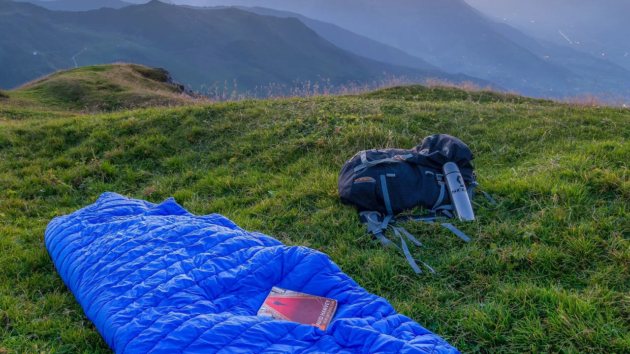 A lofted blue down sleeping bag laid out on a grassy ridge at dusk, a pack and water bottle beside it and mountain valleys behind.