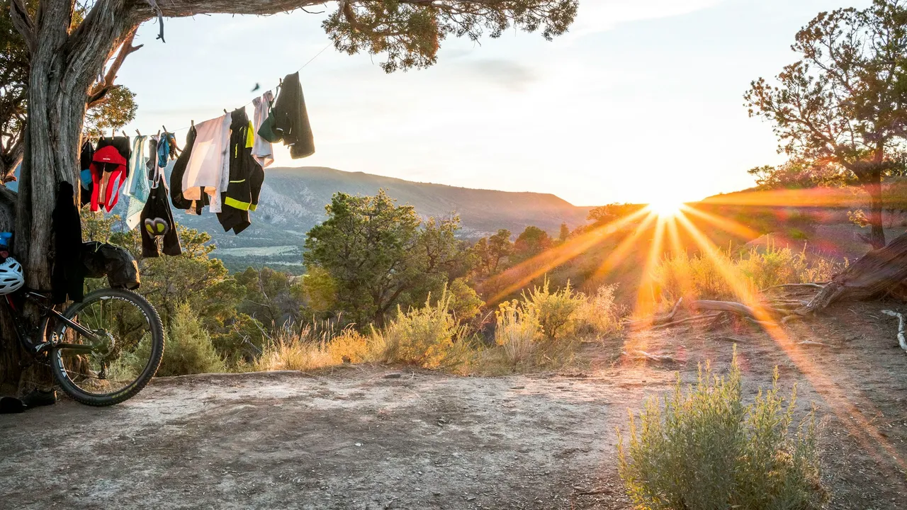 A loaded bike at a clifftop camp at sunrise, kit and clothing hung from a tree branch beside it.
