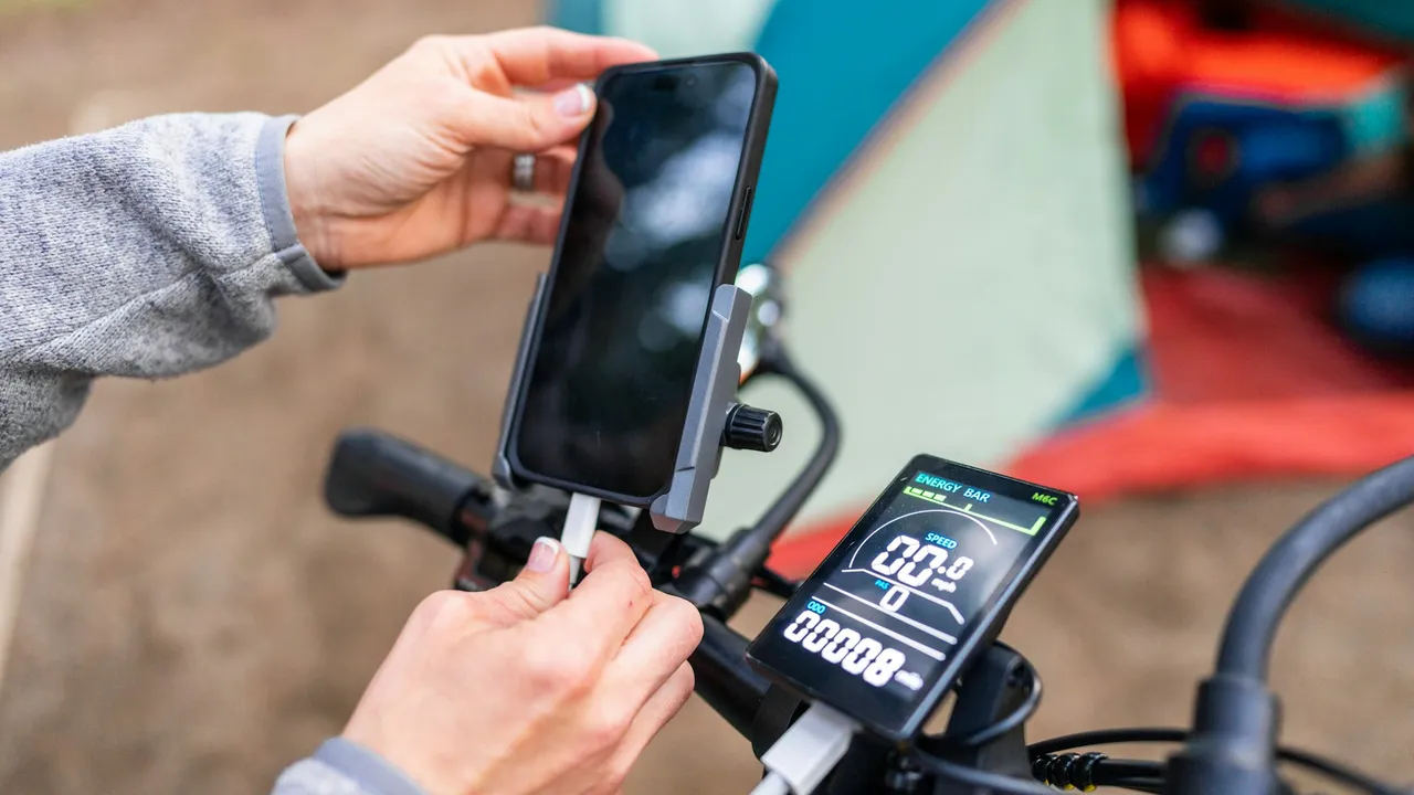 A phone mounted on a handlebar beside a bar-mounted battery display, hands adjusting the mount.