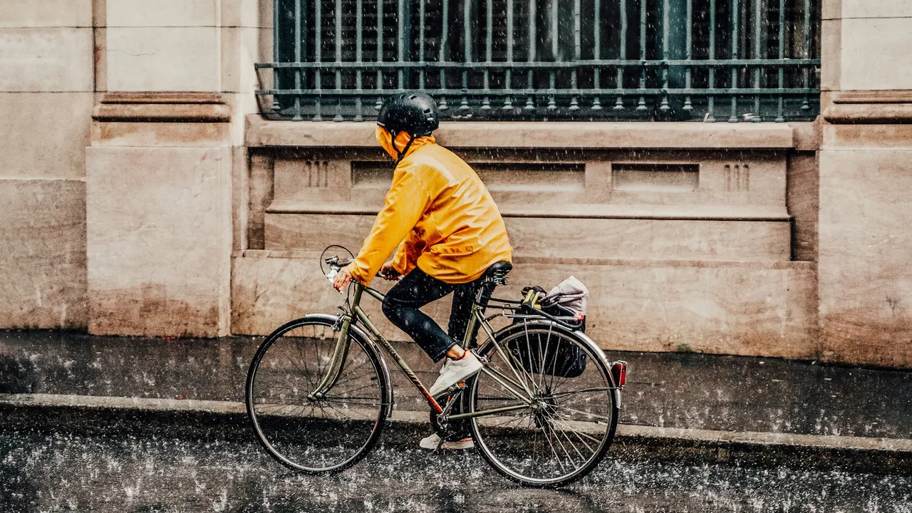 A rider in a yellow rain jacket riding past a stone building on a wet street.
