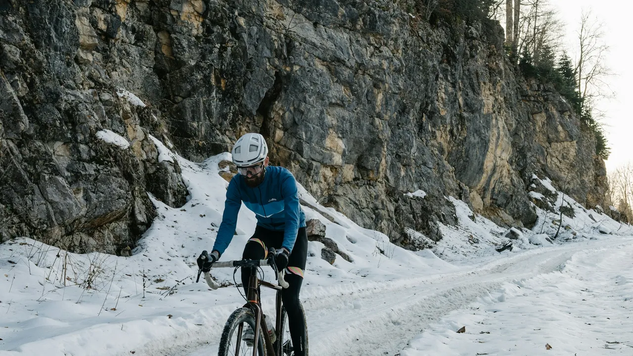 A rider on a gravel bike climbing a snow-covered road below a rock face.