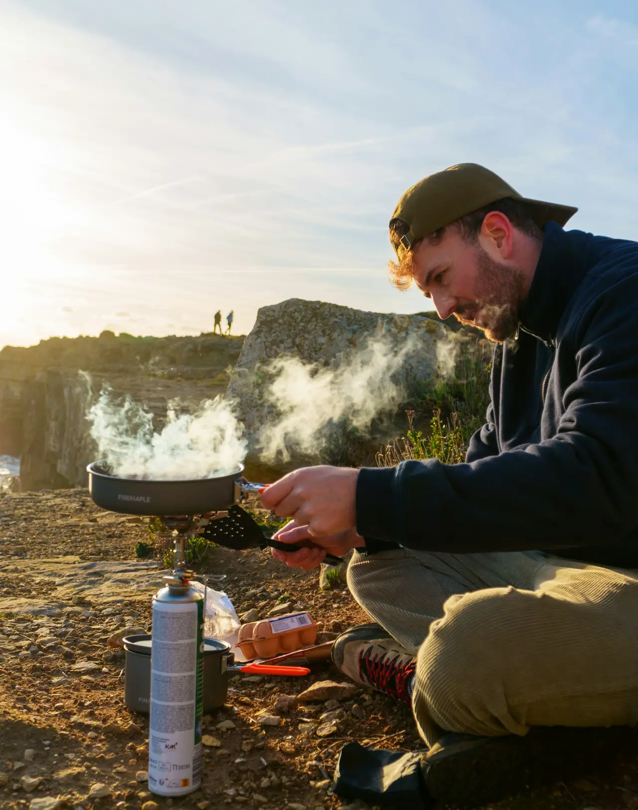 A person crouched over a camp stove at a viewpoint, steam rising from the pot.