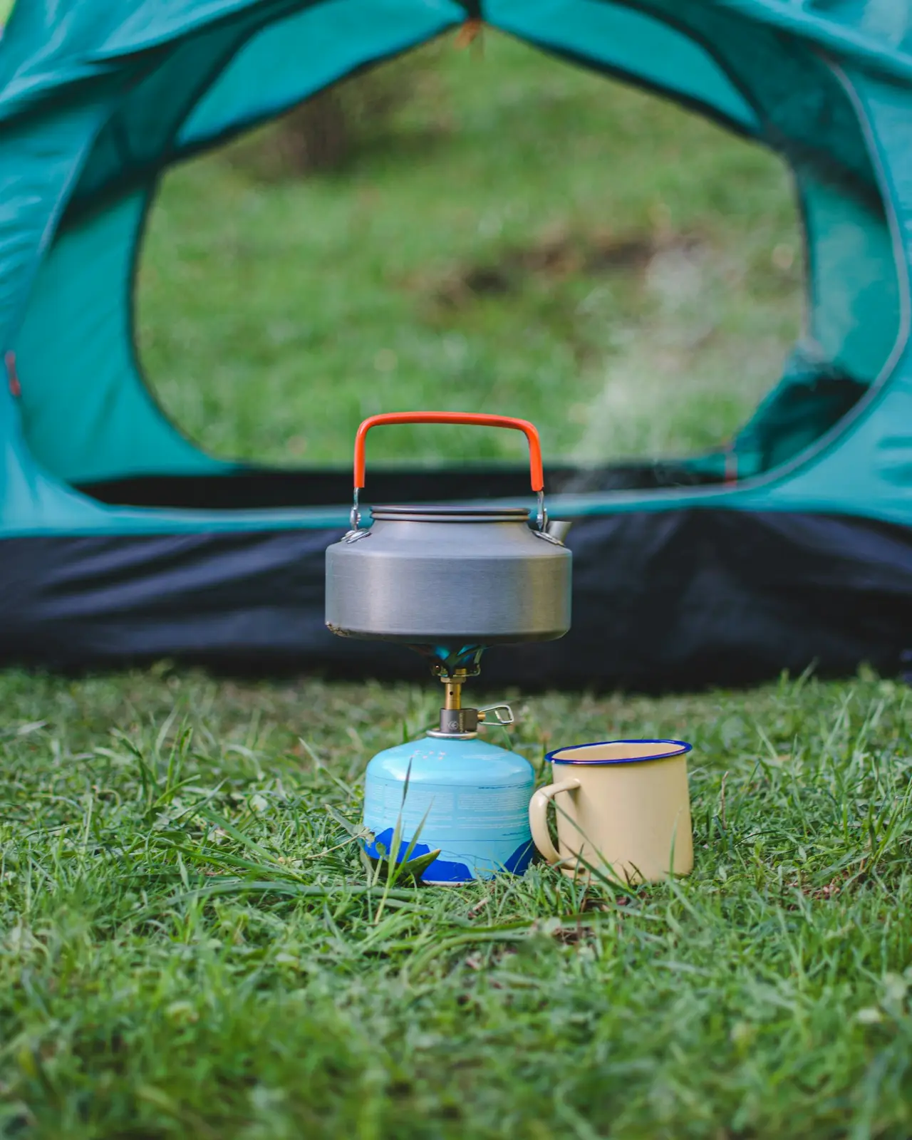 A kettle and a mug on the grass outside a pitched tent.