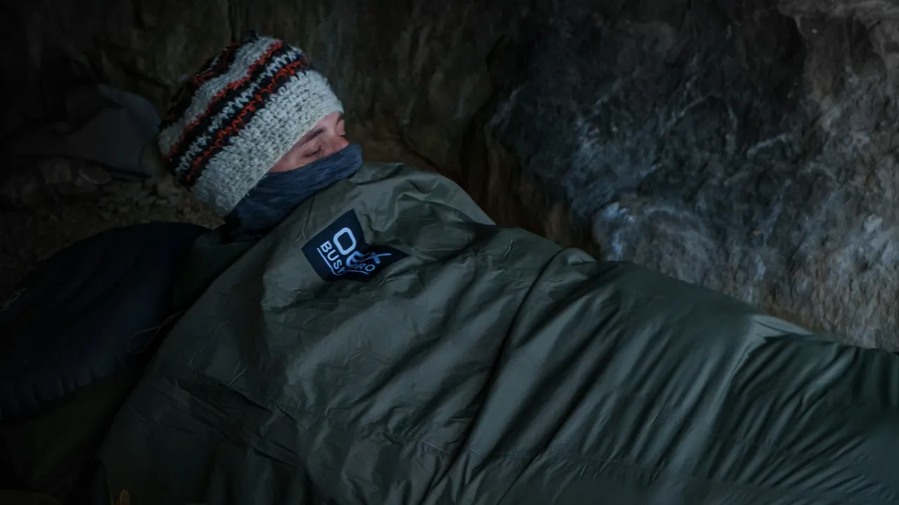 Someone in a woollen hat lying zipped into a sleeping bag under a rock overhang.