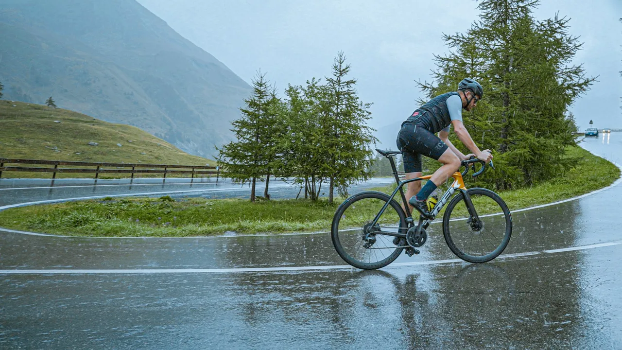 A cyclist riding a soaked mountain road, spray coming off the wheels and standing water across the tarmac.